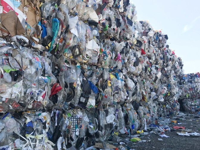 Massive piles of plastic at an outdoor recycling facility. The piles appear to be at least 20 feet him. The plastic has been compacted into tight bundles that are stacked on top of each other, then laid out in a very large L-shape. 