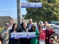 A group of people, smiling and holding thumbs up, huddle around a sign that reads "Honorary Laird Lane."