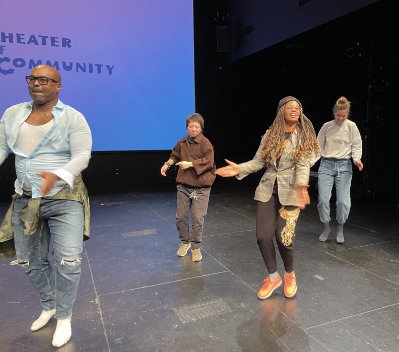 A group of people are shown dancing in a room with black walls and a screen in the rear. On that screen, the word "community" can be seen. The four dancers are dressed casually and are mid-hop. Leading the group is a woman with long, light brown braids and a dark knit cap. She is wearing black pants with a gold image on the left leg, a black T-shirt and a brown and tan plaid blazer. She's wearing orange tennis shoes.