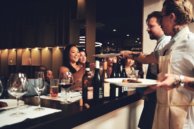 A group of people seated at a counter space that's filled with different place settings including wine bottles, and various wine glasses. A woman has her arms outstretched to receive a plate from a man wearing an apron. The rest of the seated group sits watching.  