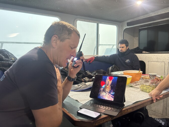 A middle-aged white man wearing a blue t-shirt and short grey/brown hair talks on a walkie talkie in the cabin of a boat. There's an ipad on a small table in front of him along with food. It's foggy through the windows of the cabin. Another white middle-aged man with black hair sits across from him wearing a blue sweatshirt, red gloves. 