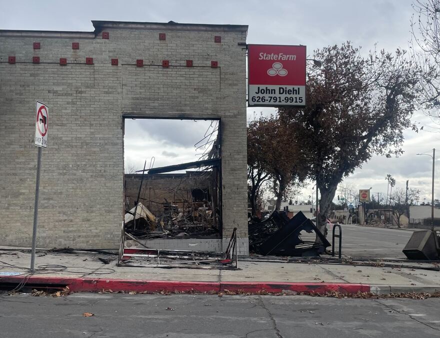 Ruins of a burned building with a State Farm sign outside. The off-white brick exterior of the building remains standing. The sign outside reads "State Farm John Diehl 626-791-9915." Wreckage of other buildings is visible in the background against gray skies.