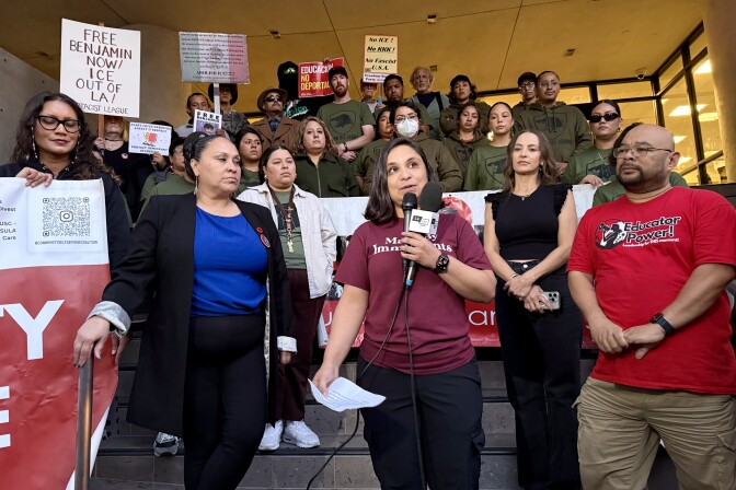 A group of people with varying skin tones. Many of them wear olive green shirts. Several people hold signs. One reads Free Benjamin Now/ ICE Out Of LA. 