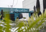 A view of Sorry Not Sorry's back outdoor patio before opening hours. There are multiple empty white tables to sit at and a teal building with the business name written on it. String lights and table umbrellas are in view with plant leaves in the foreground.