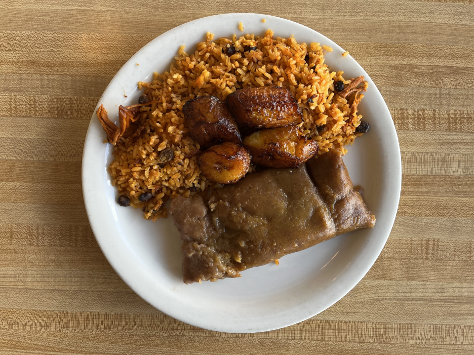 A Puerto Rican pastelle — made with green plantains, which give it a darker hue than traditional masa — sits on a white plate next to a helping of gold colored rice and beans, and fried sweet plantains. 