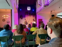 A crowd watches a woman with long light brown hair and a woman with medium-length dark hair on stage. They are both white, and most of the crowd appears to be light-skinned as well. The space features warm lighting, a fan above the stage, and metal ductwork.