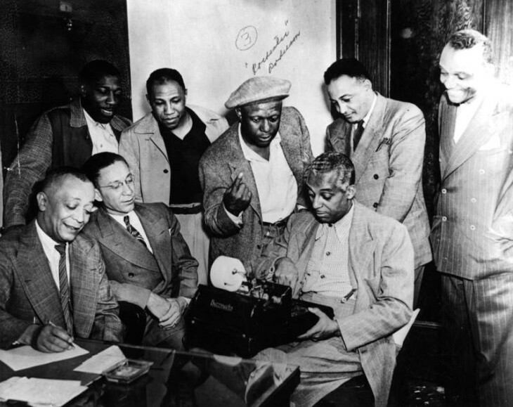 A group of men in suit jackets face a man holding an adding machine.