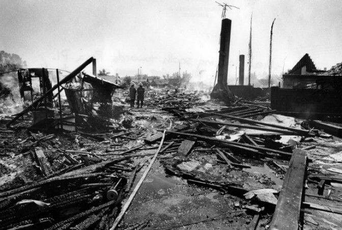 Two firemen stand in the middle of smoking plane wreckage, charred timbers and solitary chimneys with TV antennas, all that remain after a collision between an Aeroméxico jetliner and a smaller craft over Cerritos.