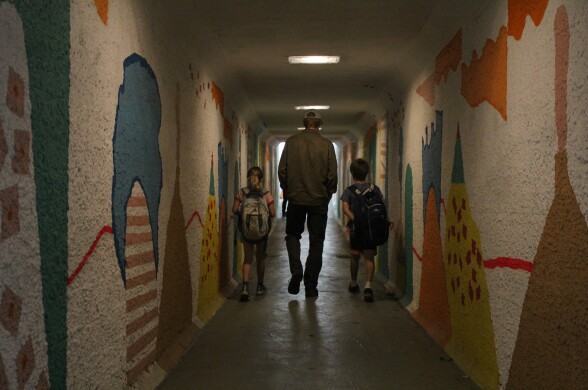 A man and two small children walk in an underground passage with art decorating the walls.