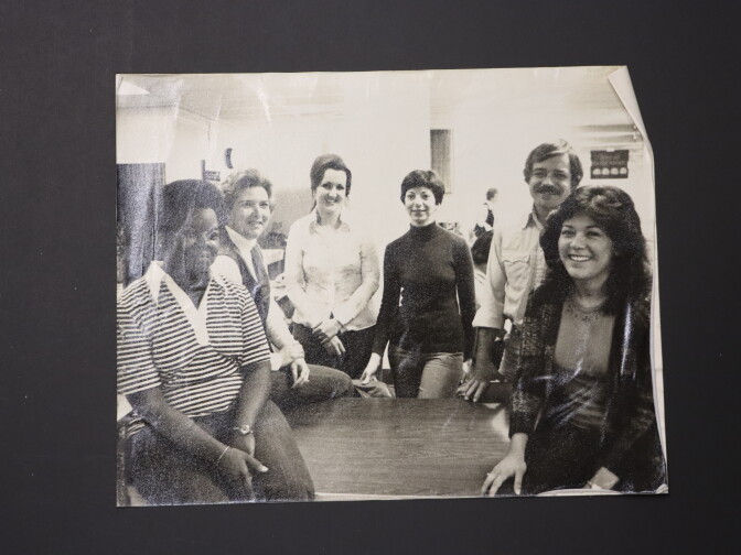 A black and white photo of five women and a man posing around a wooden table. They're all smiling and looking toward the camera.