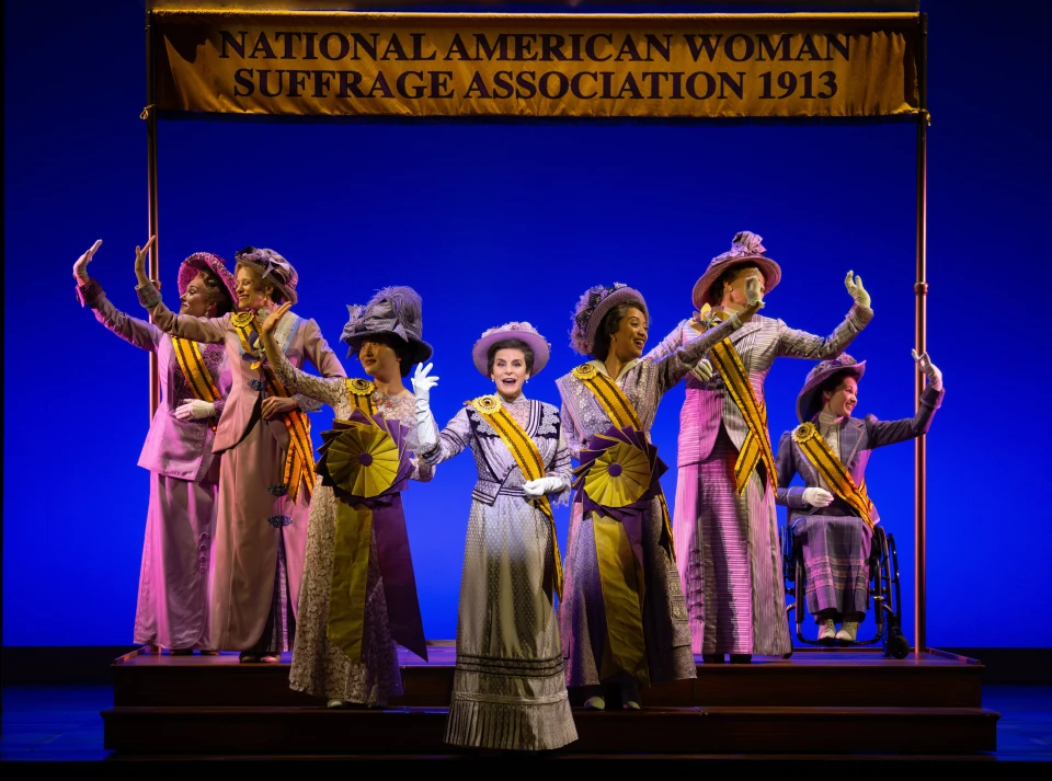 A light-skinned woman in suffragette clothing performs onstage, flanked by three women to her left and three women to her right.
