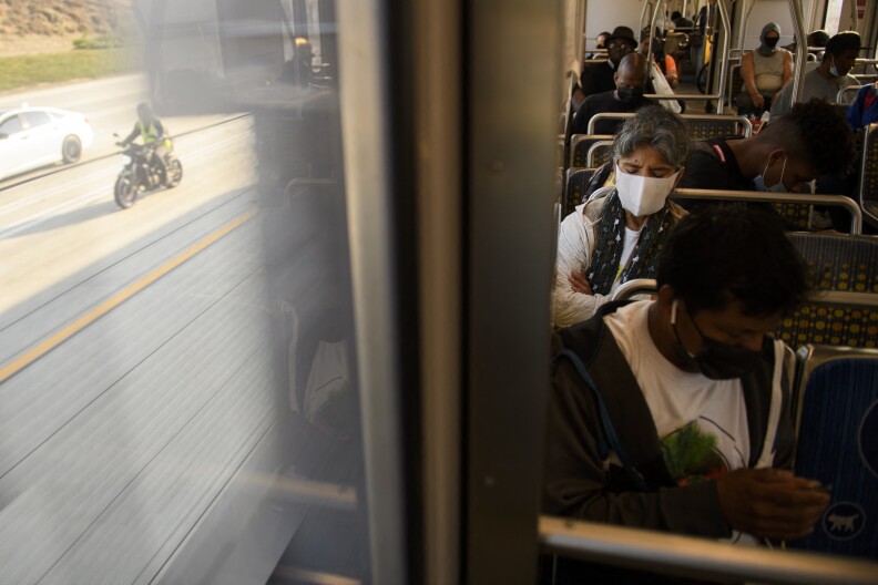 Masked public transit riders sit on a train to the right, while the window to the left shows a motorcyclist riding next to the train on a freeway.