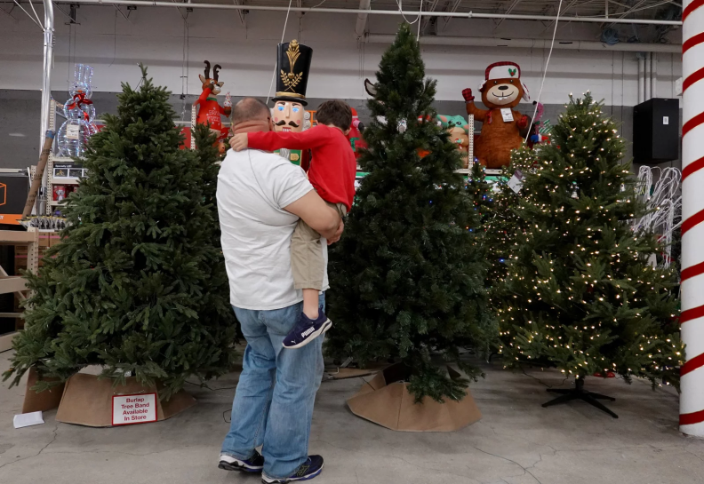 In this photo, a man who's holding a child in his arms is looking at artificial Christmas trees standing on display in a Home Depot store in Miami in 2021. The man's and child's backs are to the camera.