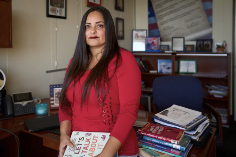 A woman with light brown skin tone dressed in red stands in front of a desk in her office that has stacks of books on it. She is holding a book in her hands, at her side.