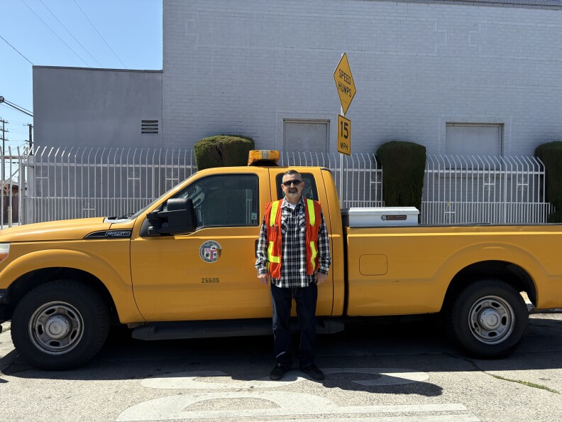 A man in a flannel and bright orange high visibility vest stands in front of a yellow truck on the road. 