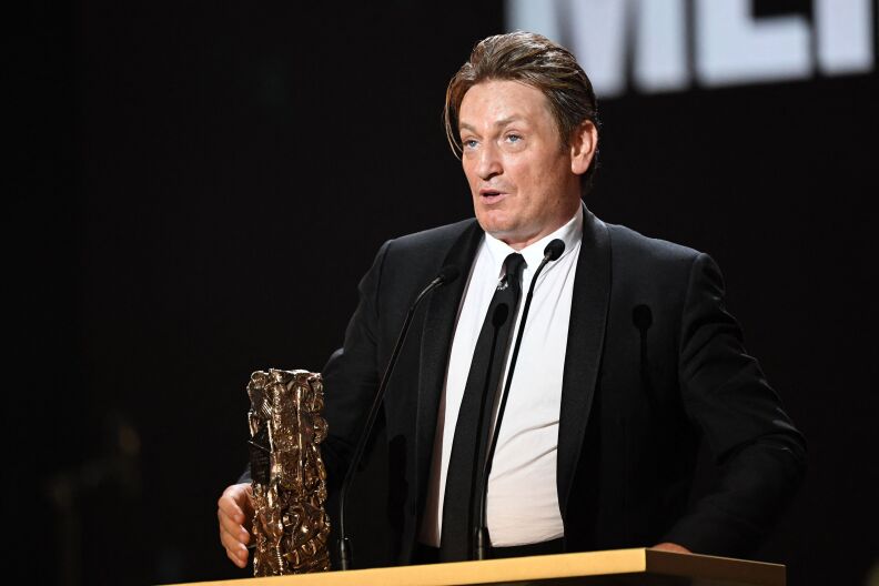 A light skinned man wearing a black tuxedo jacket, black tie and white shirt holds a gold colored Cesar award, the French equivalent of an Oscar