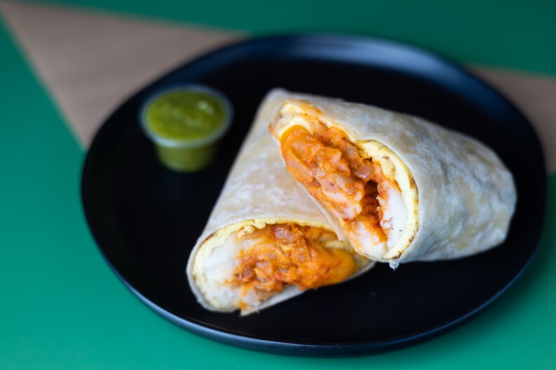 A breakfast burrito sits on a black plate. A thin layer of yellow scrambled egg is underneath a layer of white mashed potato and red kimchi, wrapped in a flour tortilla. Next to the burrito is a small container of green salsa.