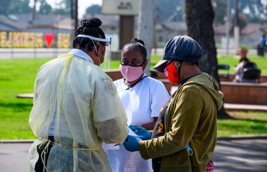 A health care workers wearing a thin yellow protective smock, a blue mask and a face shield helps two women, one wearing a pink face mask and the other wearing a blue cap and red mask, fill out paperwork at an outdoor COVID-19 mobile testing station in a parking lot.