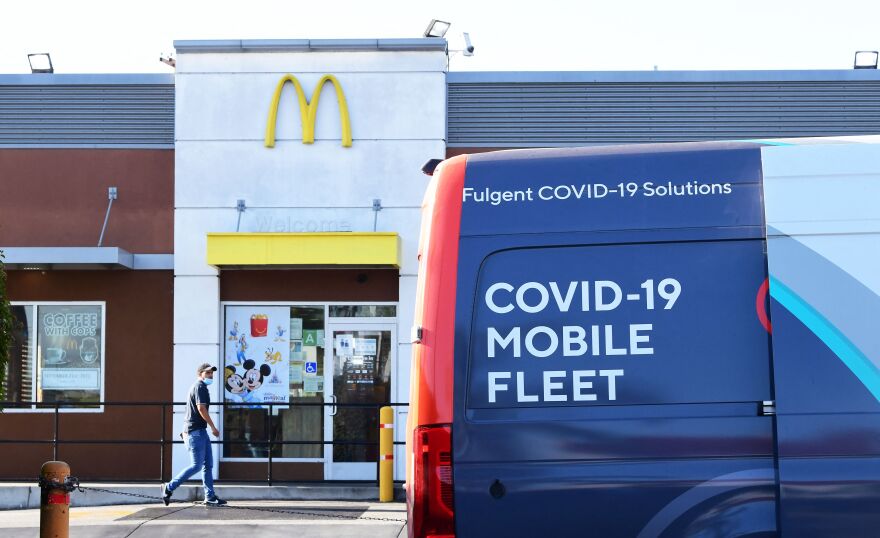 A man walks by in the background in front of a McDonald's storefront as a van with the words "COVID-19 mobile fleet" is parked on the right.