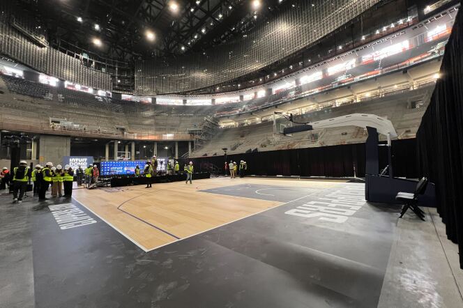A basketball arena under construction. In the background, people wearing reflective vests and hard hats are visible as well as a blue stage. 