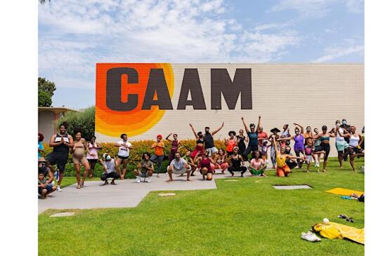 A happy group of  more than 20 African Americans standing and stretching in front of a white wall with the word CAAM in black letters 