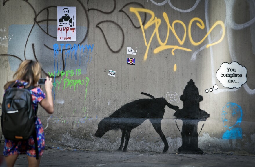 Graffiti by the secretive British artist Banksy, featuring a dog and a fire plug, draws attention on 24th Street, near Sixth Avenue in New York, on Friday, Oct. 4, 2013.  Banksy graffiti is turning up on the streets of the city and all over social media.  Banksy announced on his website that he is undertaking “an artists residency on the streets of New York” this month. 