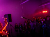 The band Saints of Sinners plays on stage under the glow of orange lights. The guitarist has long hair and is shirtless. 