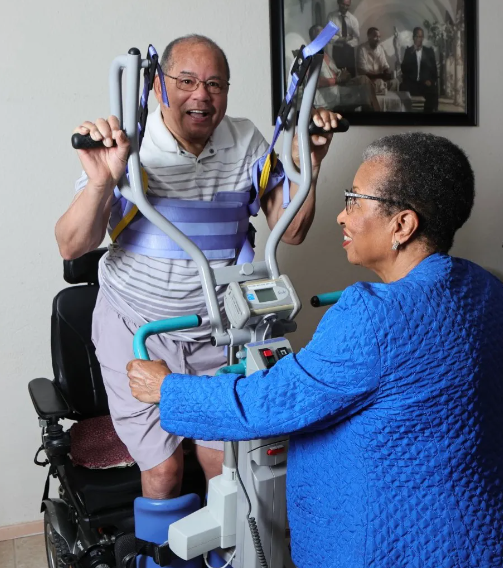 Two people with medium dark skin tone are photographed. One is a woman in a blue jacket, smiling and looking at her husband. The other is her husband, who is wearing a stripped collared shirt with knee-length cream shorts. He is smiling at the camera while using some sort of medical device where he stands and holds onto bars near his head. His wife is holding onto the device's other set of bars, near his hips.