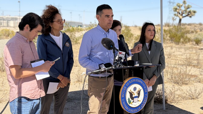 A man speaks at a House of Representatives podium outdoors in the desert, with four others standing nearby under a tent.