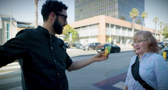 A man with a light skin tone and dark hair, wearing glasses and a black chef's coat, extends a colorful taco toward a woman with light hair and a light skin tone on a sunlit Los Angeles sidewalk. The woman, wearing a plaid shirt with a crossbody bag, smiles mid-conversation as office buildings and palm trees line the street behind them.