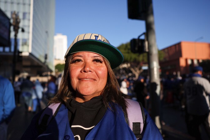 A woman with medium-light skin tone wearing a white and blue striped Dodgers hat stands in the sunlight for a portrait. 
