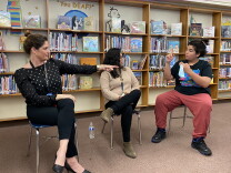 An 8th grader wearing a black t-shirt, reddish pants and black shoes sits in a chair close to two adult women. The women watch the 8th grader's hands are mid-motion in front of his face as he communicates in American Sign Language.