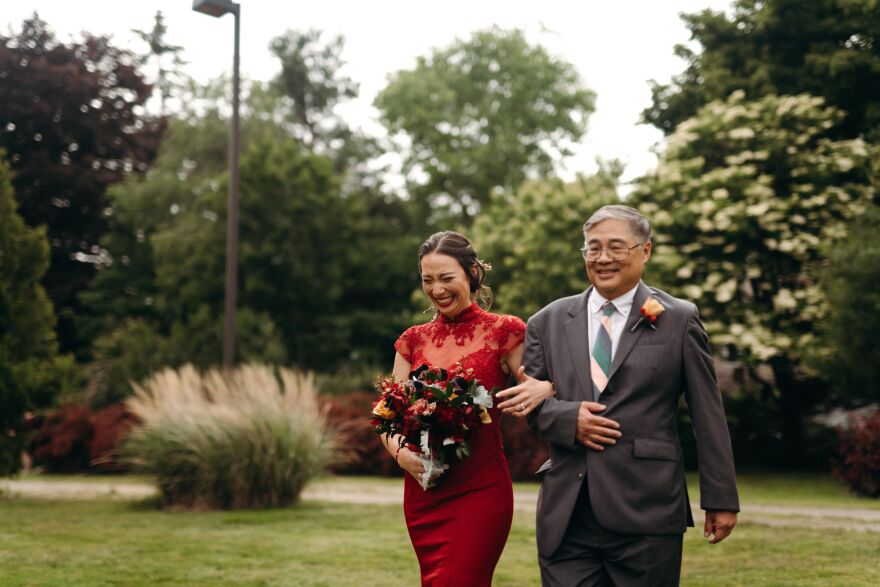 An Asian woman in a red dress holds a bouquet of flowers and walks arm in arm with an Asian man wearing glasses and a grey suit, in a garden.