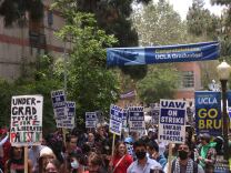 People holding signs that read "UAW ON STRIKE" march on a campus near a banner that says "Congrats Graduates!"
