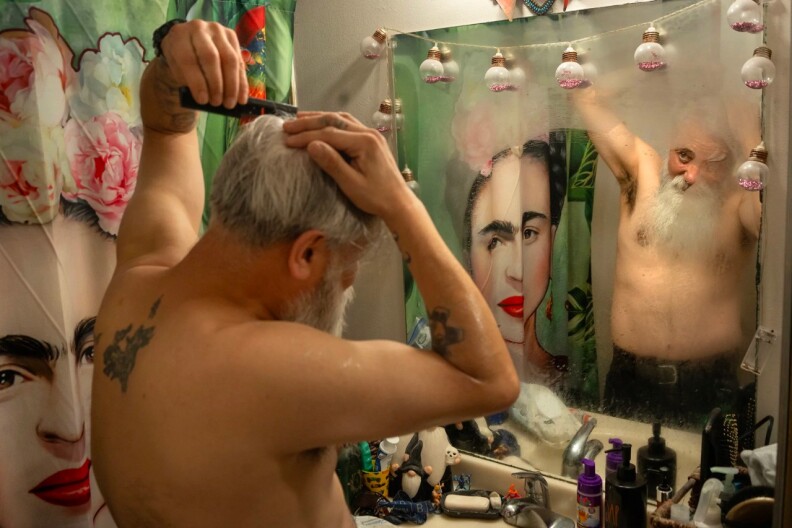 An shirtless older man with a large gray beard and a tattoo on his back combs his hair while looking at himself in a foggy mirror.