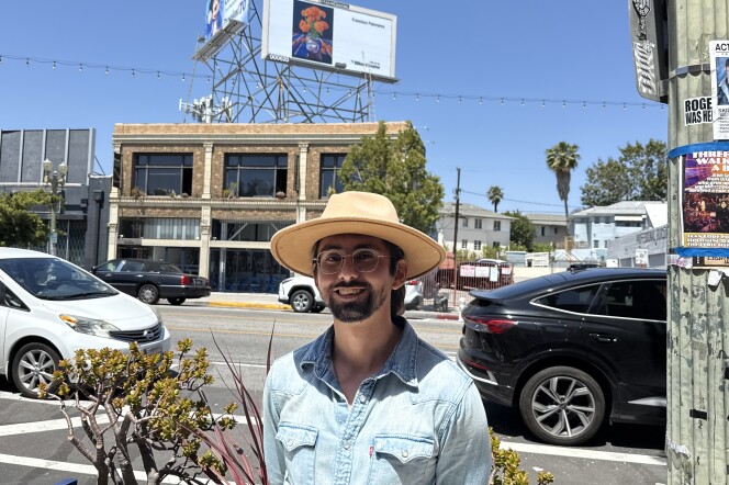 Artist Francisco Palomares looks at the camera. He wears glasses and a large brimmed hat. Behind him is a billboard depicting his oil painting "Fabuloso." The painting depicts a bouquet of orange marigolds in a purple Fabuloso bottle. 