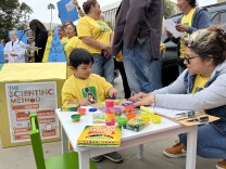 A 4-year-old boy with light brown skin plays with playdough at a children's table set up as part of a rally. People hold signs, chanting behind him. 