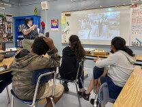 Three students in a public school classroom sit facing away from the camera, their eyes pointed to their teacher, wearing a floral-print button down shirt at the front of the classroom. The teacher points at a projection on her classroom's white board of a black-and-white archival photo, depicting a man in a tuxedo and two women in dresses.
