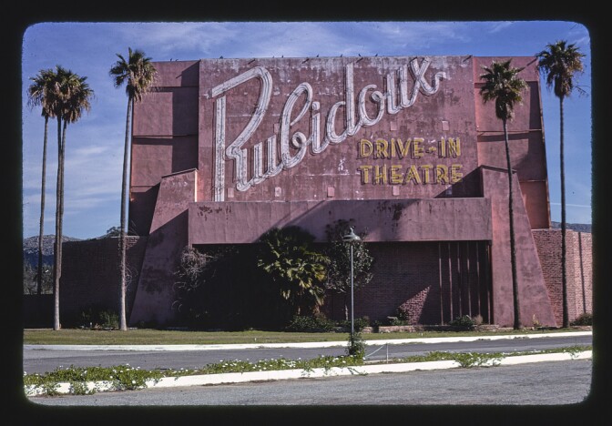 The words Rubidoux Drive-In Theatre in unlit neon appears on a wall flanked by tall palm trees