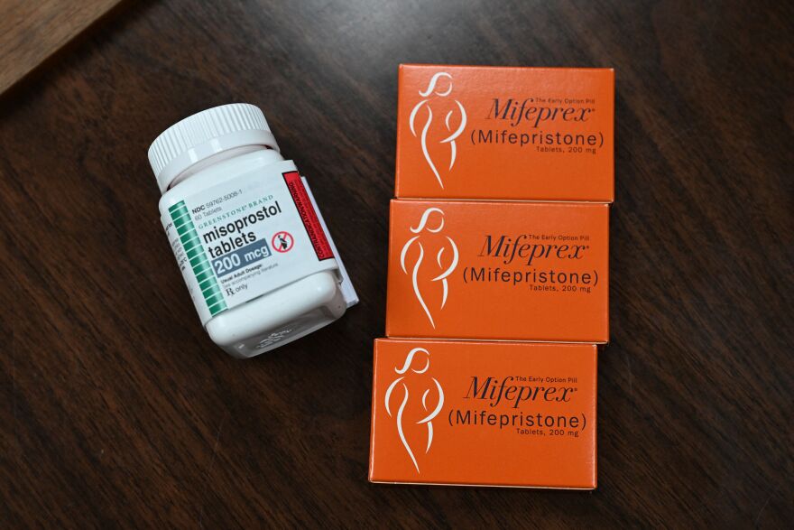 A white pill bottle is labeled misoprostol tablets next to three tomato red containers with the figure of a woman and the words Mifeprex (mifeprisone)