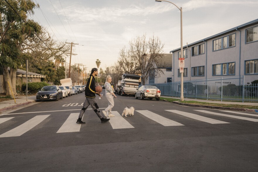 A Black man wearing gray sweats and a black and yellow sweater holds a basketball as he crosses the street on a crosswalk with a white blond woman in gray sweats holding a leash with a small white dog.