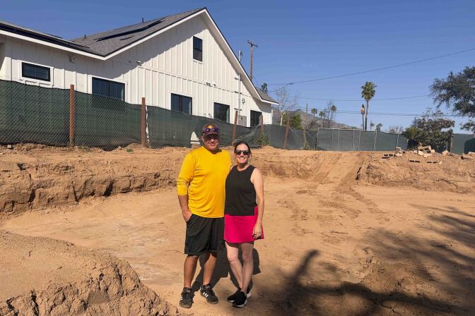 Two people stand in a lot of a property where the ground has been dug a few feet down. The new home next to them is separated with a green tarp and chain link fence.