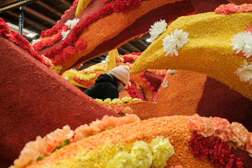 A float decorator stands in the middle of a towering parade float covered with yellow, orange and red flowers in a Pasadena warehouse.
