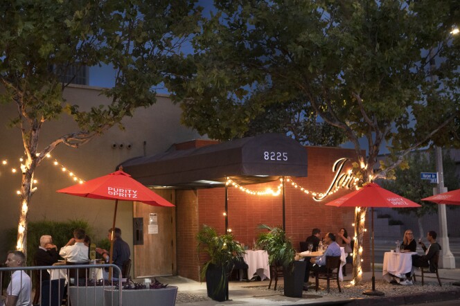 An image of the exterior of Jar, a restaurant located in the Beverly Grove neighborhood of Los Angeles.  The outdoor dining area consists of various tables and chairs underneath red umbrellas, all positioned around the restaurant's front entrance, which is covered by a long black awing next to a red brick wall.
