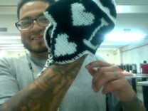 A bearded man in a t-shirt and glasses smiles as he holds up a crocheted black-and-white cap with two hearts on the side.