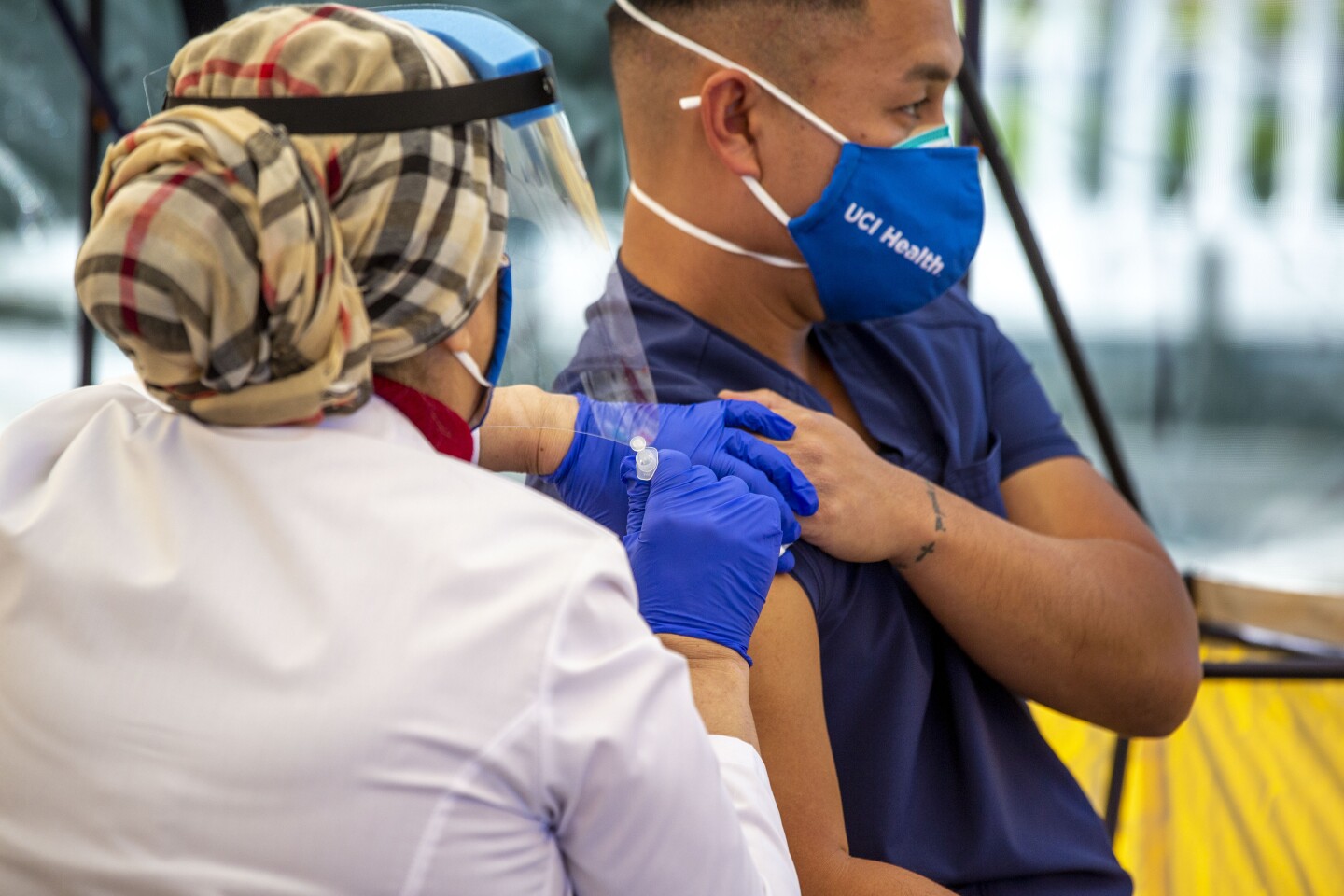 A health care worker in blue gloves and a mask and face shield uses a needle to innoculate a man wearing blue scrubs and a UCI Health face mask.