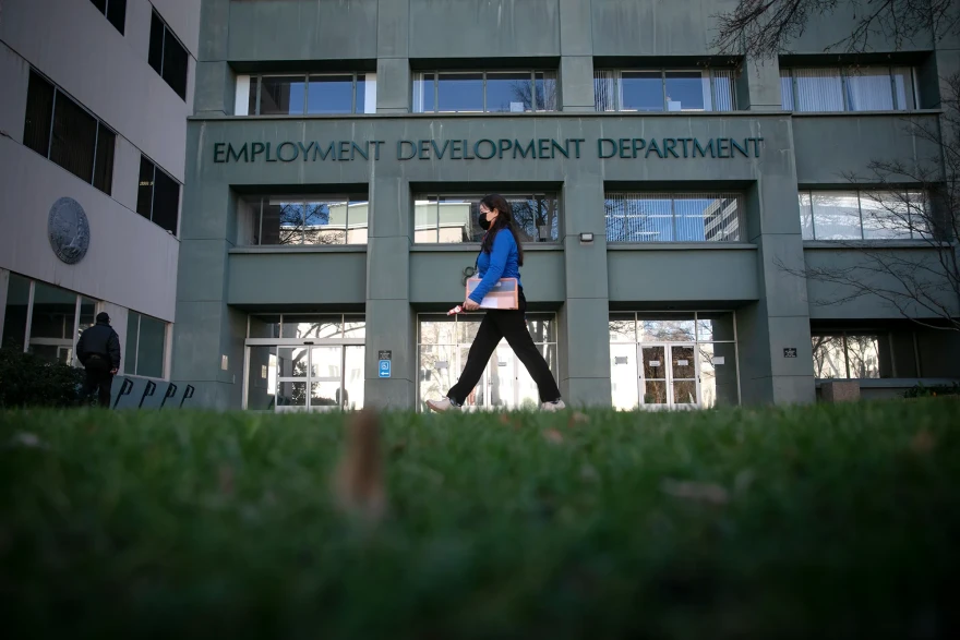 A woman wearing a blue long sleeved top and black pants walks past a large, dark green building with signage that reads, "Employment Development Department"