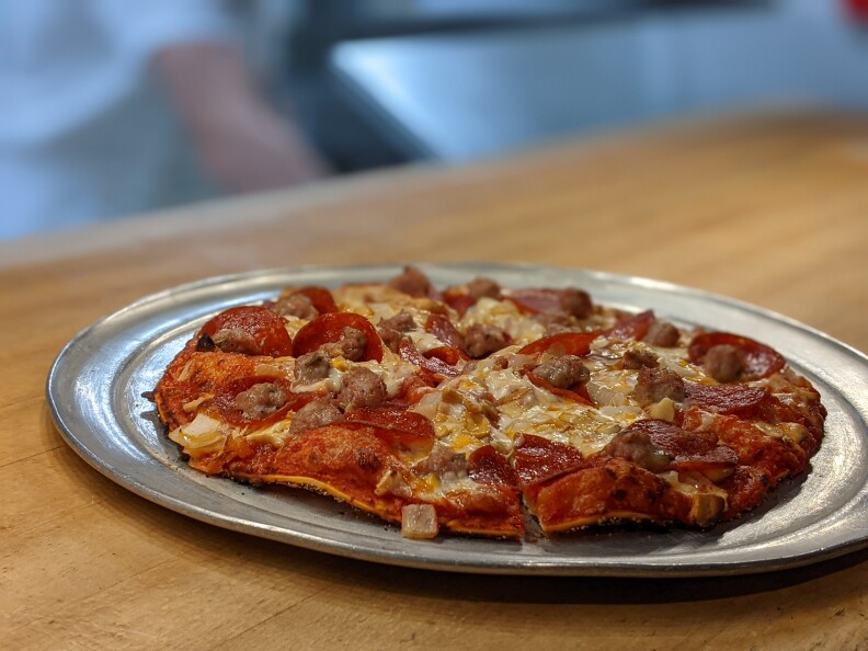 A color image of pizza sitting on a round metal tray. The tray itself sits on a light brown table top.