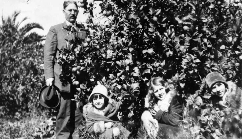 A black and white photo of a family in a lemon grove; an older light skinned man is standing to the side, wearing glasses and a 1920's suit and tie; he is holding his hat. Next to him three light skinned children crouch down and look off to the side