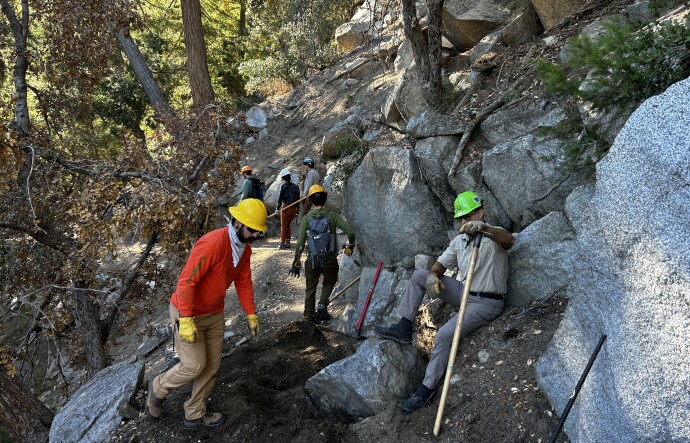 Multiple younger and middle-aged men on a dirt trail with large boulders wearing hardhats and holding McLeod tools do work on a trail. 